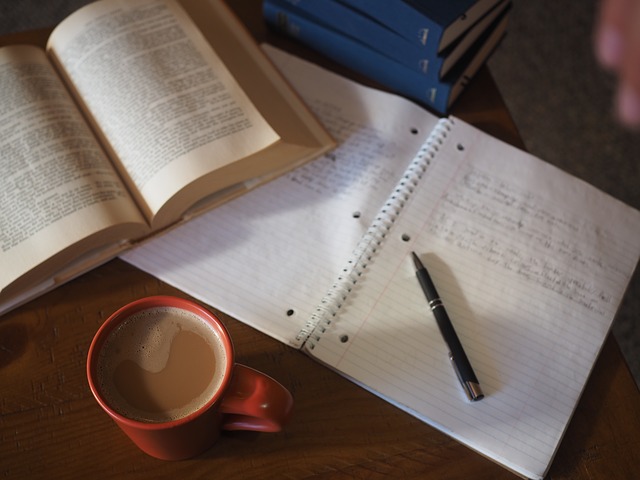Imagen de una zona de estudio compuesto por un libreo abierto, una libreta con apuntes, una taza de café y algunos libros.
